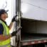 Commercial technician performing a box truck roll-up door repair inspection by checking the lift cables, rollers, and track at a loading dock before a summer delivery route.